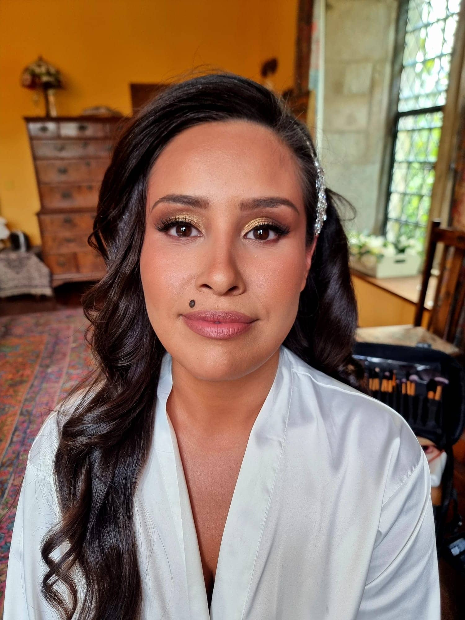 ana-nettlestead-place-bridal-hairstyle-elegant-updo.jpg A woman with long, wavy dark hair styled in Bilsington Priory bridal hair and make-up sits indoors, wearing a white dressing gown and natural make-up with a bold beauty mark above her lip. A window, furniture, and yellow walls are visible in the background.