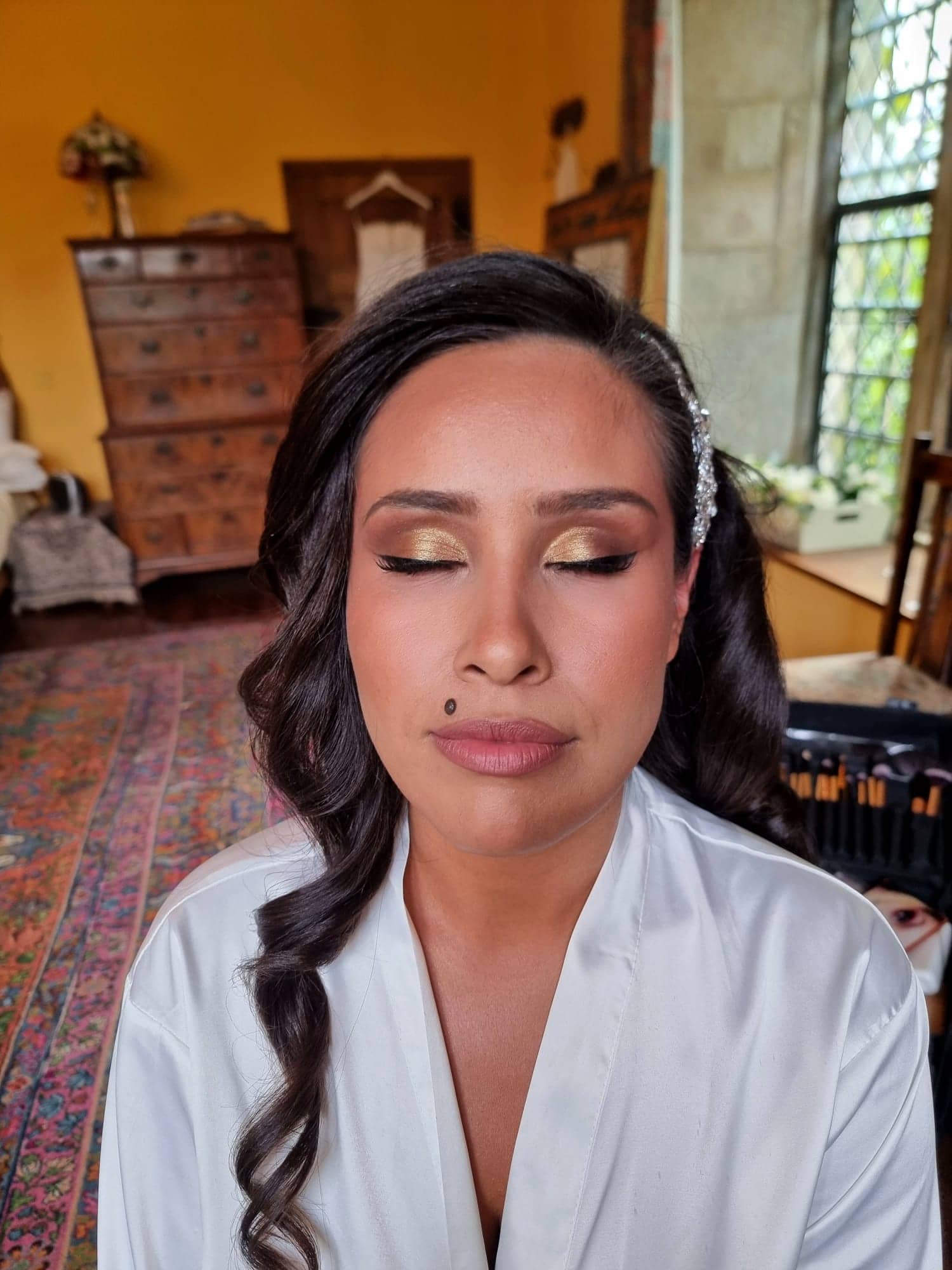 ana-nettlestead-place-bridal-preparation-wedding-morning-kent.jpg A woman with long dark hair and gold eyeshadow, styled for Bilsington Priory bridal hair and make-up, sits with her eyes closed in a warmly lit room filled with vintage furniture, a colourful rug, and soft natural light streaming through the window.