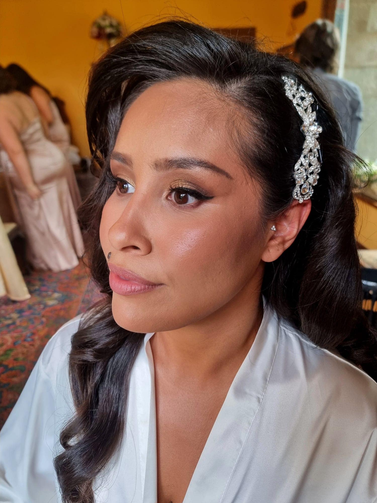 ana-nettlestead-place-bride-garden-venue-kent.jpg A woman with long dark hair styled in waves and adorned with a jewelled hairpiece looks to the side. She wears a white satin dressing gown and showcases Bilsington Priory bridal hair and makeup. Blurred figures and warm tones are visible in the background.