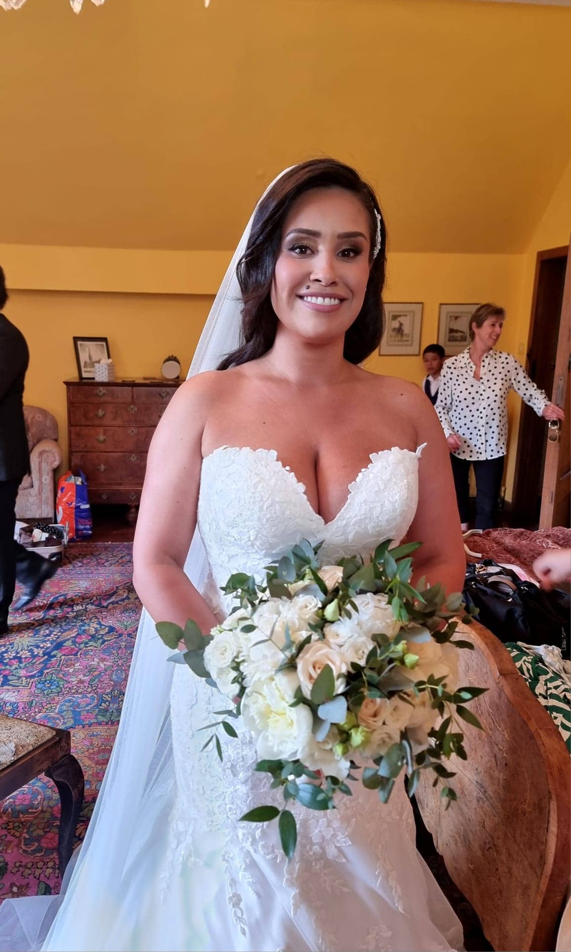 ana-nettlestead-place-bride-portrait-kent.jpg A smiling bride in a white lace wedding dress, with elegant Bilsington Priory bridal hair and make-up, holds a bouquet of white flowers and greenery. She stands indoors with people and vintage furniture in the background.