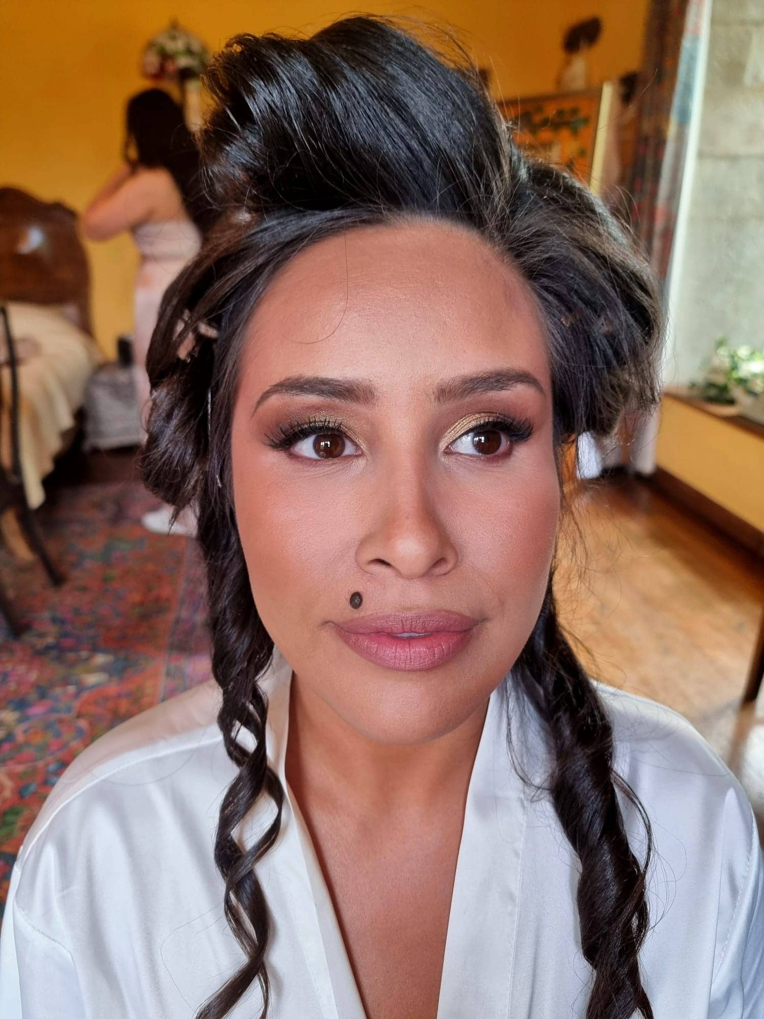 ana-nettlestead-place-wedding-bridal-hair-makeup.jpg A woman with styled curly hair and make-up sits indoors wearing a white dressing gown. The background shows another person near a bed in a warmly lit room with wooden floors and yellow walls, capturing the elegance of Bilsington Priory bridal hair and make-up.