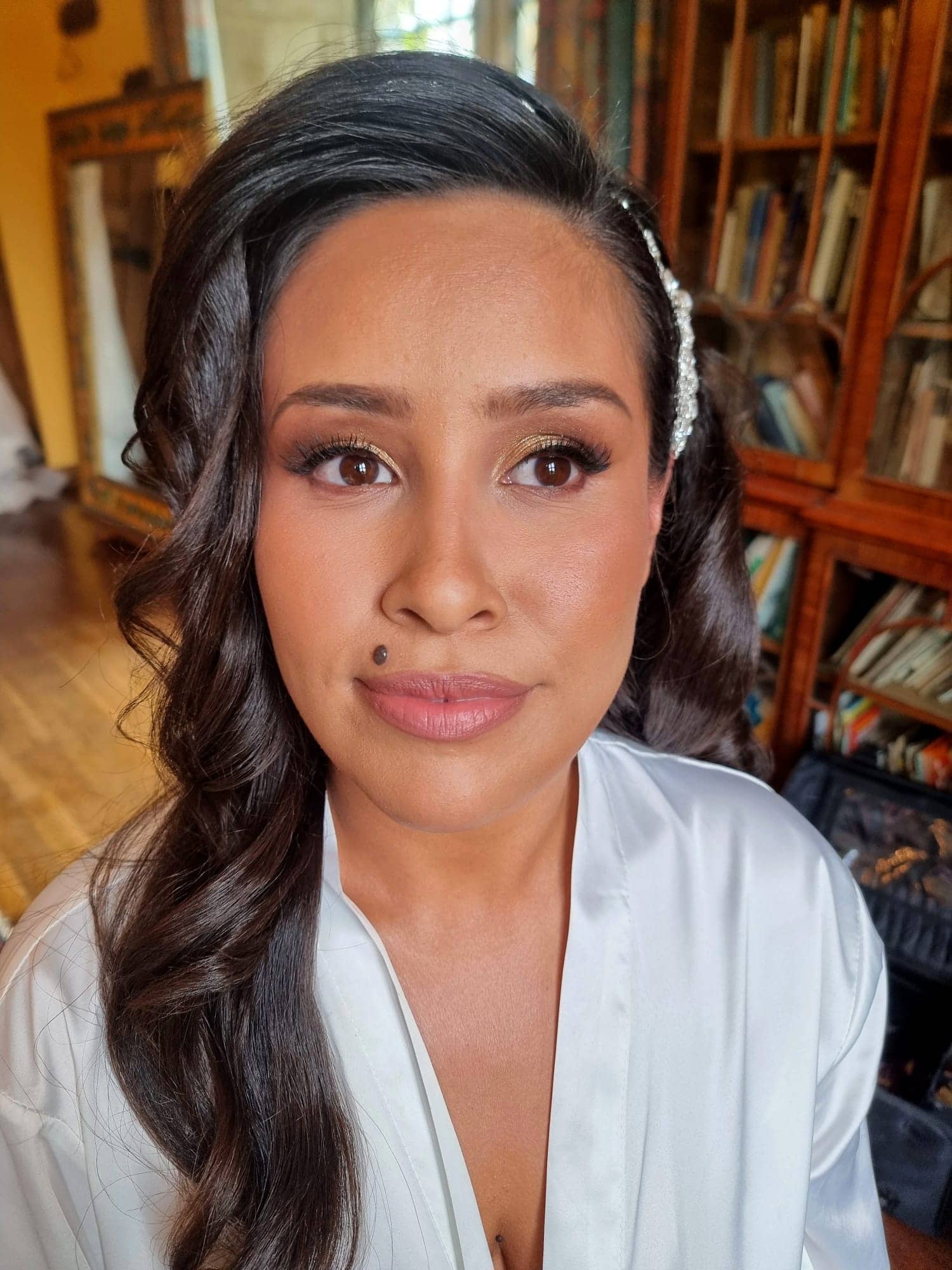 ana-nettlestead-place-wedding-makeup-natural-glow.jpg A woman with long, wavy dark hair and a white dressing gown sits indoors, showcasing elegant Bilsington Priory bridal hair and make-up. She has natural make-up, a beauty mark above her lip, and a decorative accessory. Bookcases and a wooden floor are visible behind her.