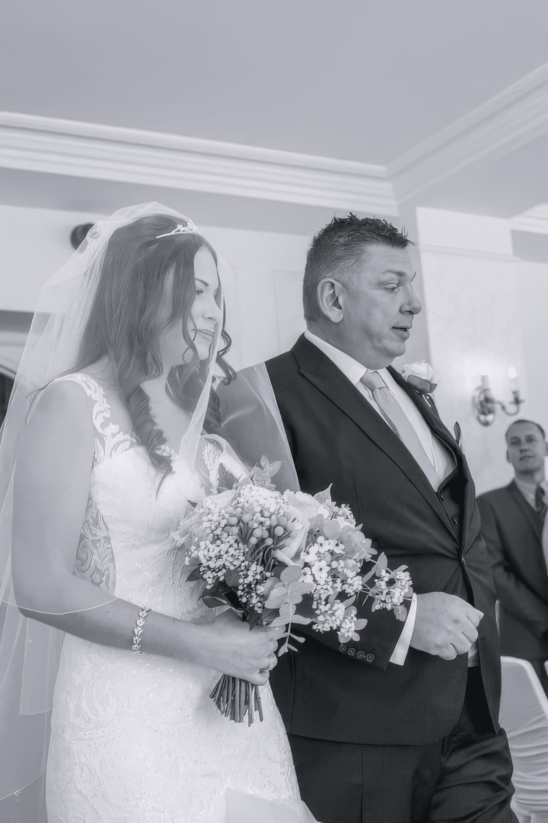 gabi-hythe-imperial-bridal-look-natural-glow.jpg A bride in a white dress and veil holds a bouquet whilst standing beside a man in a suit, possibly her father, during a wedding ceremony. Another man in a suit is seen in the background.