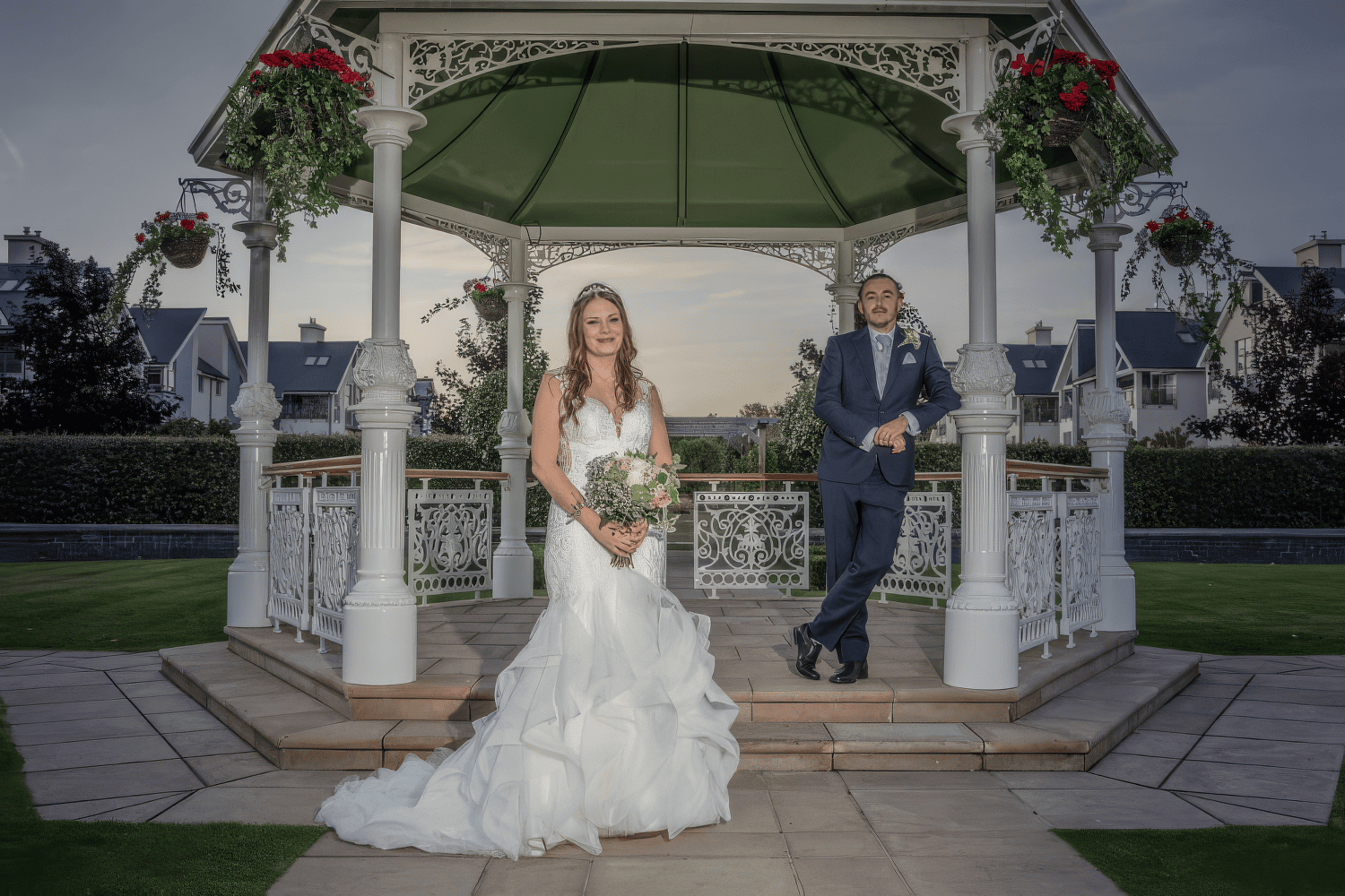 gabi-hythe-imperial-bride-coastal-venue-kent.jpg A bride in a white dress holds a bouquet and stands in front of a groom in a suit, who leans against a white gazebo decorated with hanging flower baskets in a garden.