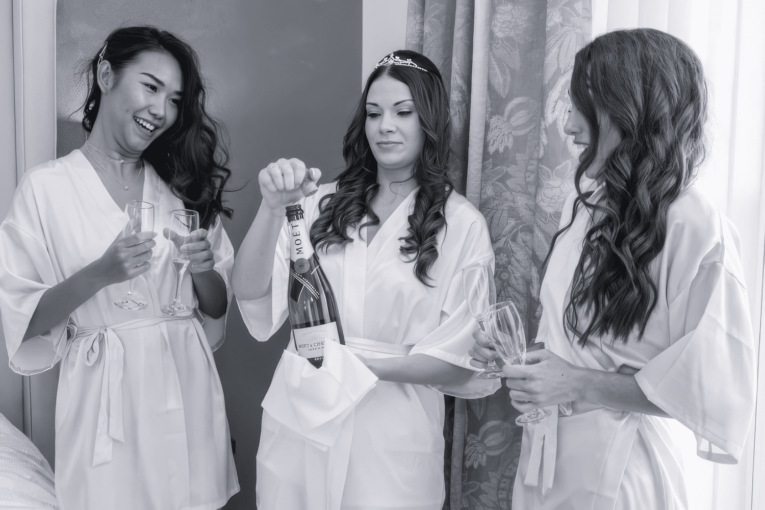 gabi-hythe-imperial-bride-elegant-finish-kent.jpg Three women in dressing gowns stand together, holding champagne glasses. The woman in the centre opens a bottle of Moët & Chandon champagne while the other two smile and laugh. All have long hair and appear to be getting ready for a celebration.