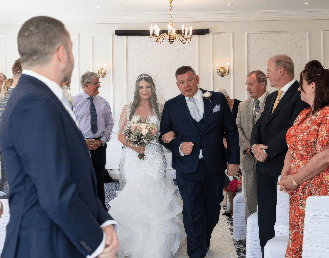 gabi-hythe-imperial-bride-hair-accessory-detail.jpg A bride in a white dress and veil walks down the aisle holding a bouquet, escorted by a man in a dark suit. Guests, seated and standing, watch as a groom waits at the front in a navy suit. The room is elegantly decorated.